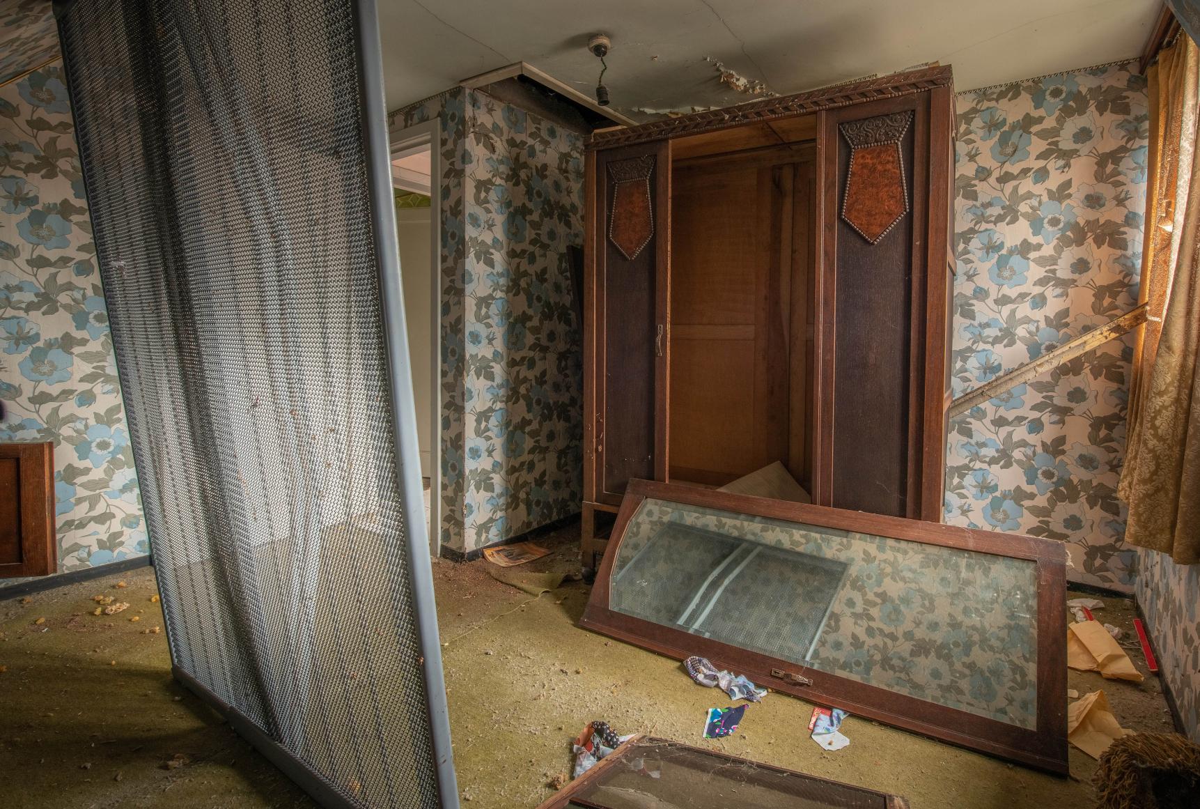 Vor der Renovierung zeigt x²o Badezimmer ein stark veraltetes Badezimmer mit auffälliger floraler Tapete in Blau- und Beigetönen, einem beschädigten Holzschrank mit offenen Türen, herumliegenden Möbelteilen und einem abgenutzten Boden, was einen unordentlichen und alten Gesamteindruck vermittelt.