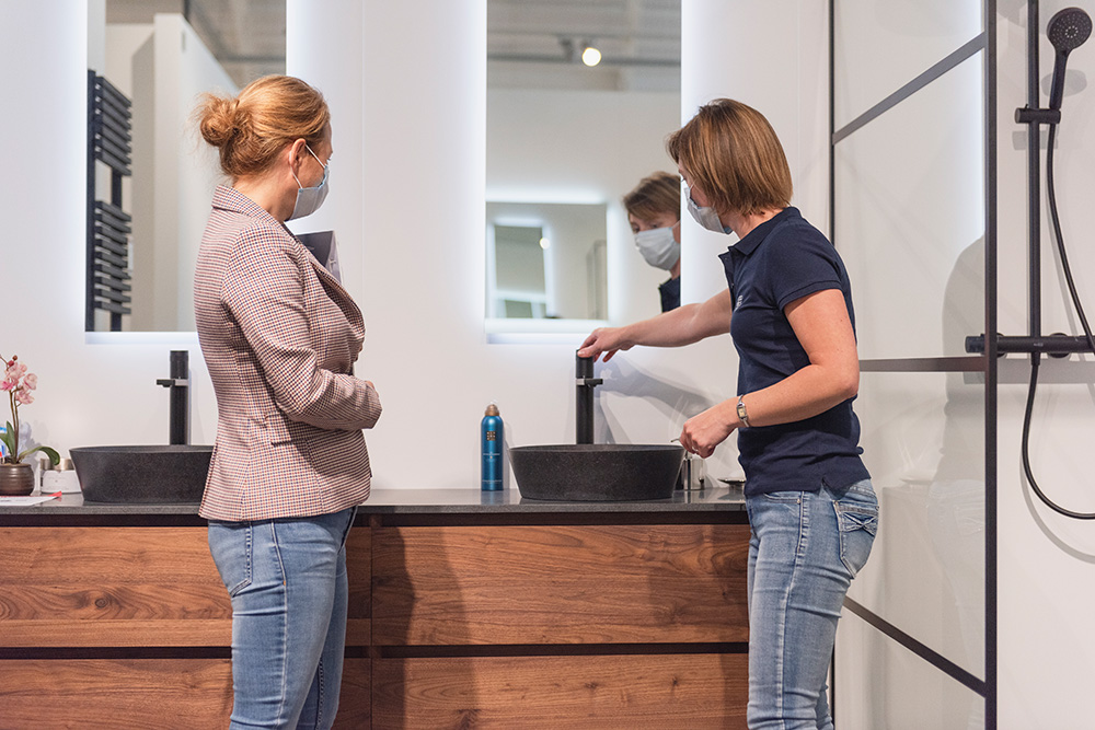 In diesem Showroom von x²o Badezimmer stehen zwei Frauen mit Mundschutz vor einem Badezimmermöbel aus Holz mit einem schwarzen Aufsatzwaschbecken. Eine der Frauen bedient den Wasserhahn, während sie gemeinsam beraten. Im Hintergrund sind Spiegel und eine schwarze Duschwand zu sehen, was die moderne und beratungsorientierte Showroom-Atmosphäre zeigt.