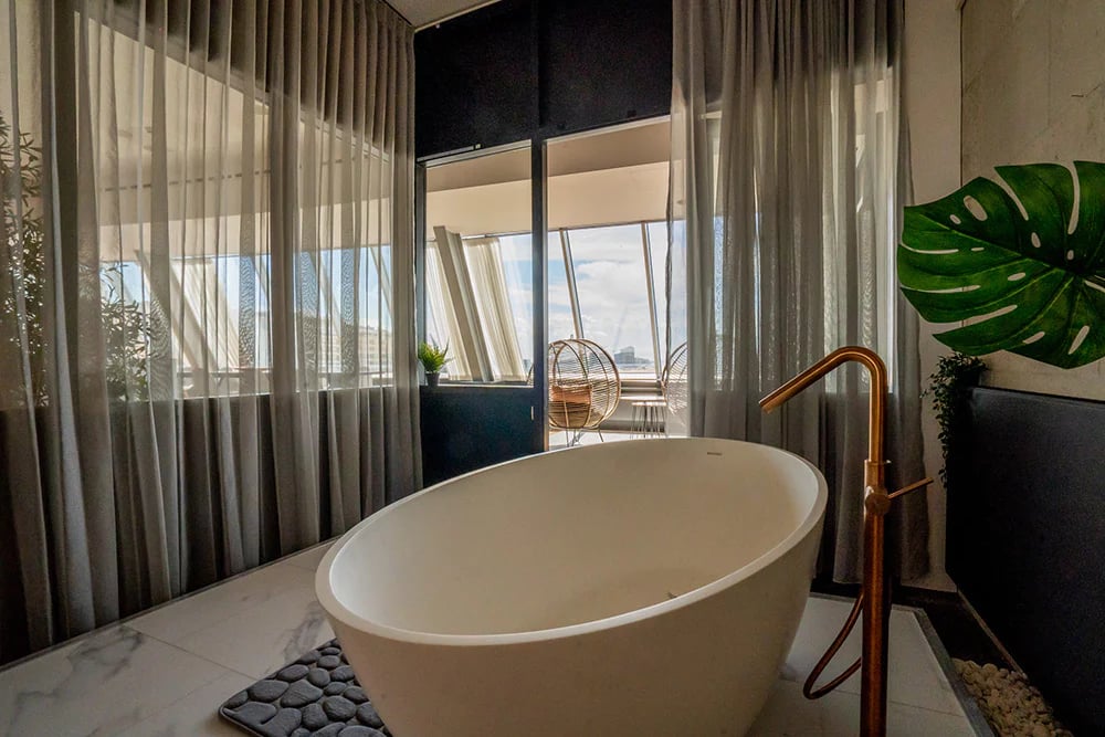 Freistehende weiße Badewanne vor großen Fenstern mit transparenten Vorhängen, kombiniert mit goldfarbener Armatur und natürlichen Akzenten in einer exklusiven x²o Badezimmer Atmosphäre.