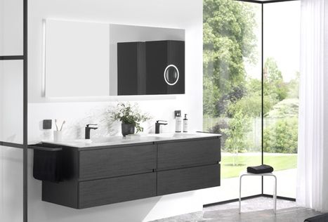 Meuble de salle de bains avec double vasque dans une configuration moderne de x²o salles de bains sous un plafond incliné. Le meuble est réalisé en bois clair avec de larges tiroirs. Deux miroirs verticaux sont fixés au mur au-dessus des vasques. Une plante décorative est visible à gauche et une baignoire avec zone douche à droite.