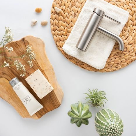 Photo à plat d’un robinet de lavabo finition inox coolmetal posé sur un plateau en bois, entouré d’accessoires de salle de bains comme des serviettes, du savon, des plantes et des éléments en pierre naturelle. Cette composition met en valeur le design des robinets de x²o salles de bains.
