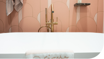 Baignoire îlot blanche de x²o salles de bains avec robinetterie dorée sur pied, installée devant un mur de carreaux roses aux motifs arrondis. Une serviette est posée sur le rebord de la baignoire, créant une ambiance bien-être.