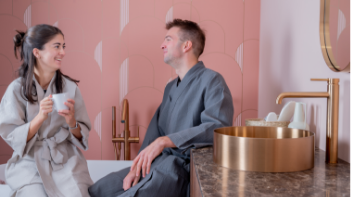 Salle de bains chaleureuse de x²o salles de bains montrant deux personnes en peignoir discutant près d’une baignoire îlot blanche. Au premier plan, un lavabo rond couleur laiton posé sur un plan en marbre, avec des carreaux muraux roses et des robinetteries dorées.