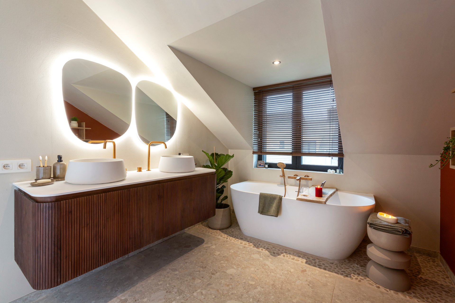 Eine stilvolle x²o Badezimmer mit freistehender Badewanne, Holzwaschtisch mit zwei Aufsatzbecken und runden Spiegeln mit Beleuchtung unter einer Dachschräge. Neben der Badewanne steht eine Pflanze und ein kleiner Steintisch mit Kerzen.