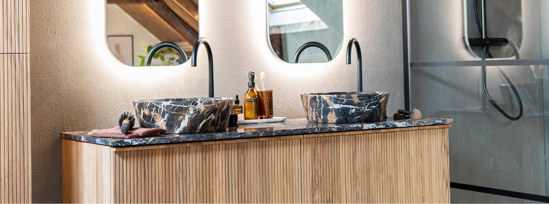 Gros plan sur une double vasque x²o badkamers avec deux lavabos ronds en marbre foncé, des robinets noirs et un meuble en bois à rainures verticales. Des produits de soin, une brosse à dents et une brosse sont posés sur le plan. En arrière-plan, deux miroirs éclairés et une cabine de douche vitrée avec détails noirs.