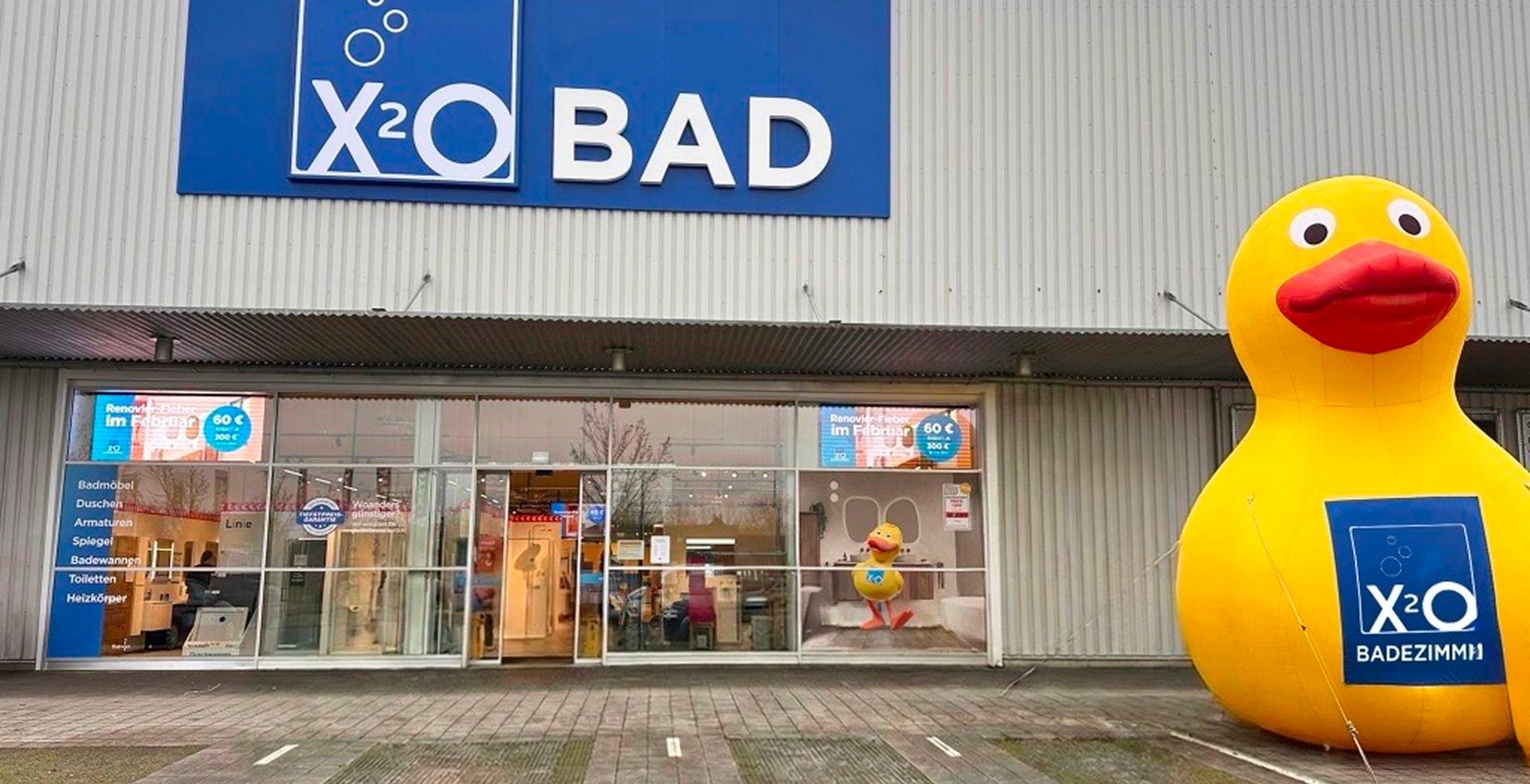 Fassade des x²o Badezimmer Showroom in Mannheim mit großem blauem X²O BAD-Logo über dem Eingang und einer aufblasbaren gelben Ente mit X²O-Schild neben dem Gebäude.