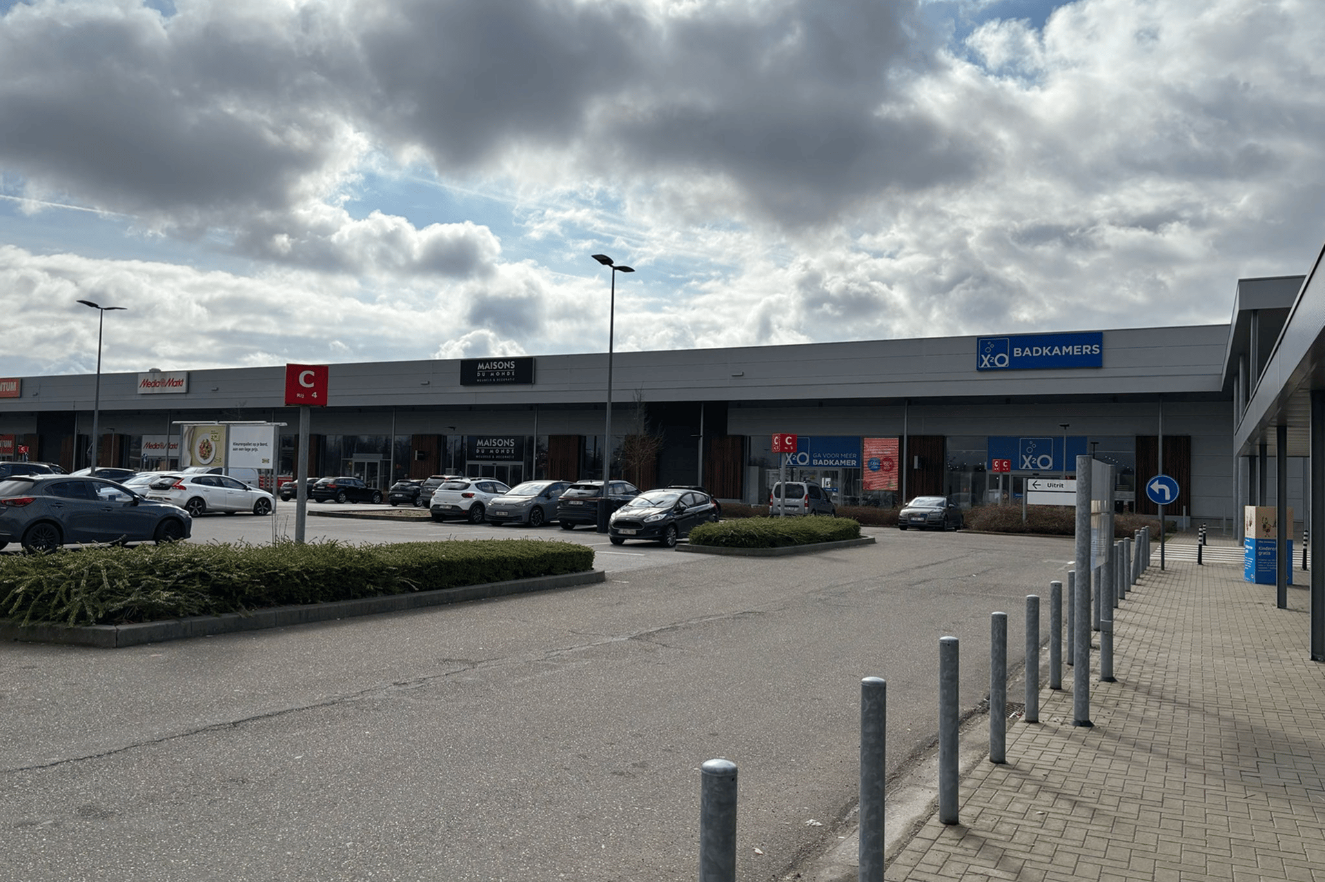 Centre commercial avec plusieurs magasins, dont XO Salles de Bains, et des voitures garées au premier plan sous un ciel nuageux.