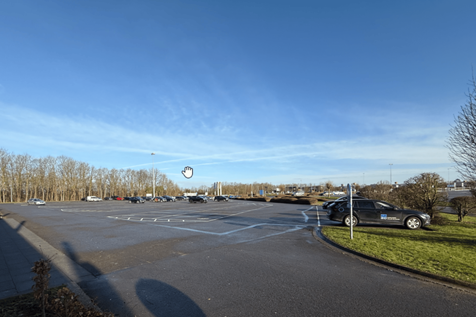 Grand parking presque vide avec quelques voitures sur les côtés, sous un ciel bleu clair et des arbres en arrière-plan.