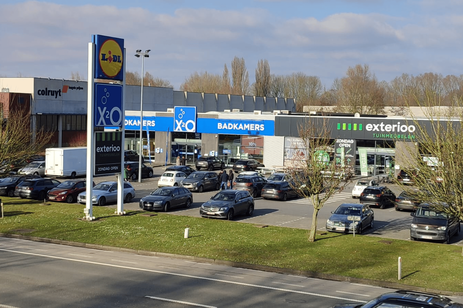 Centre commercial avec XO Salles de Bains, Exterioo et d'autres magasins, un parking bien rempli au premier plan et un panneau Lidl le long de la route.