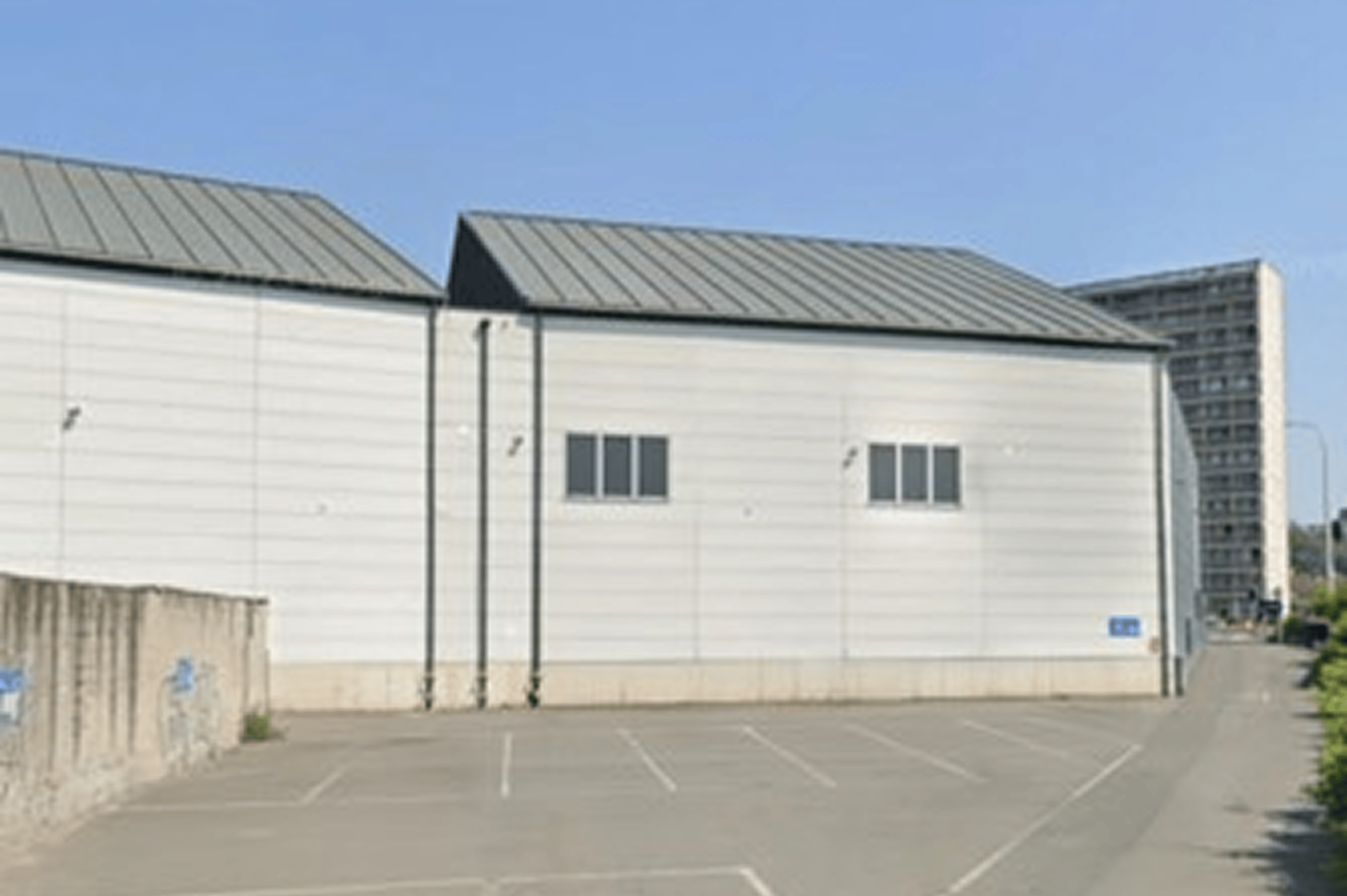 Zijaanzicht van een wit industrieel gebouw met een metalen dak, enkele ramen en lege parkeerplaatsen op een zonnige dag.