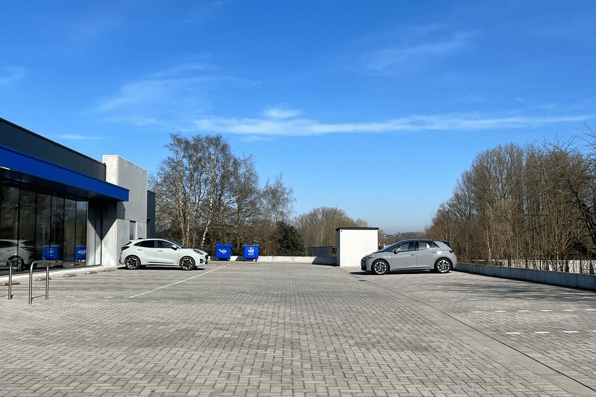 Grand parking presque vide avec quelques voitures et des conteneurs bleus, situé à côté d’un bâtiment à façade vitrée sous un ciel bleu clair.