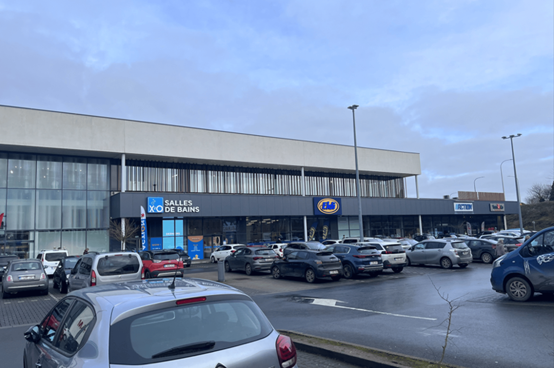 Centre commercial avec X2O Salles de Bains, JBC et Action, devant un parking très fréquenté par une journée nuageuse.