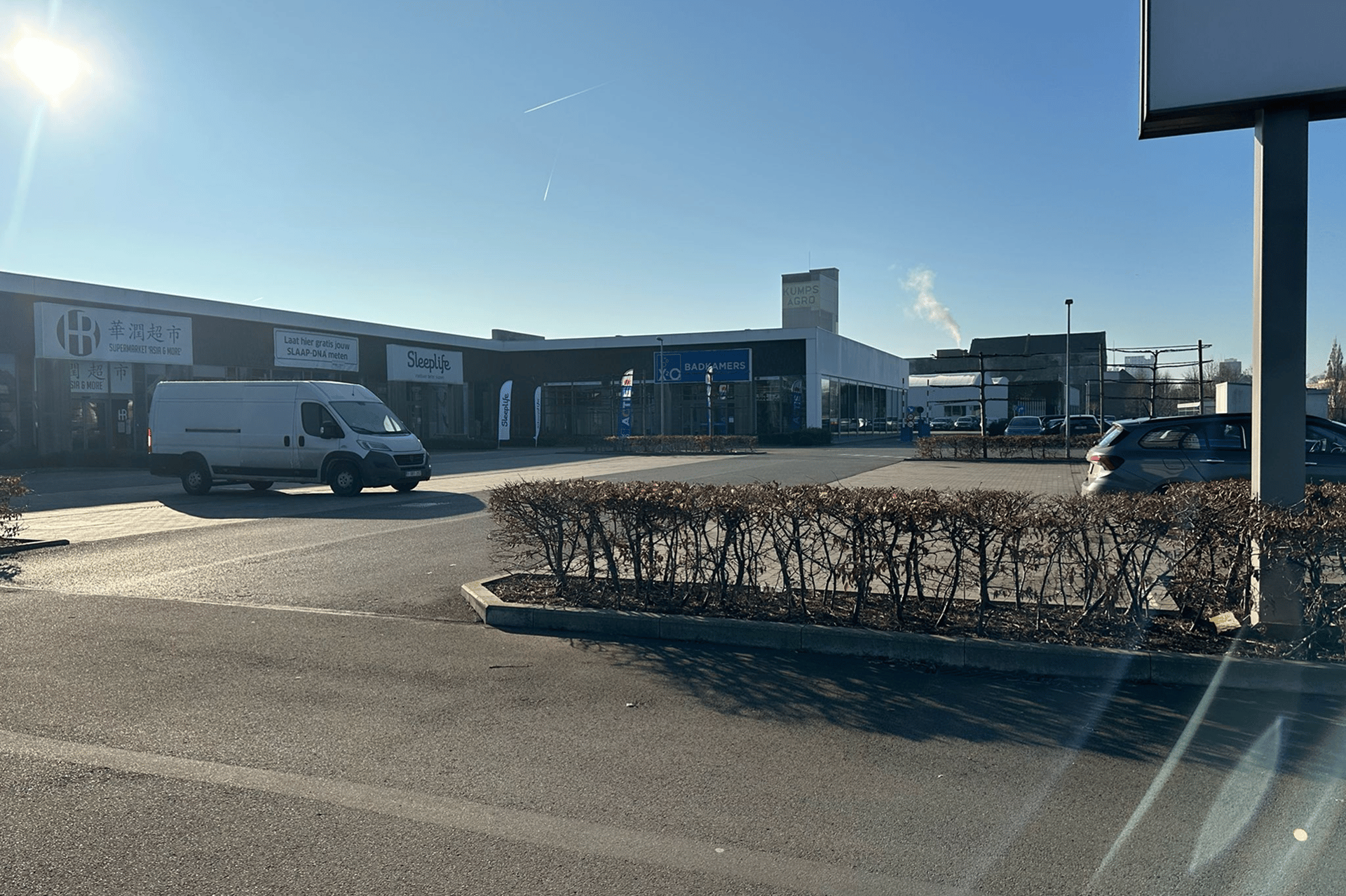 Centre commercial avec X2O Salles de Bains et d’autres magasins, avec une camionnette et des voitures sur le parking sous un soleil éclatant et un ciel bleu.