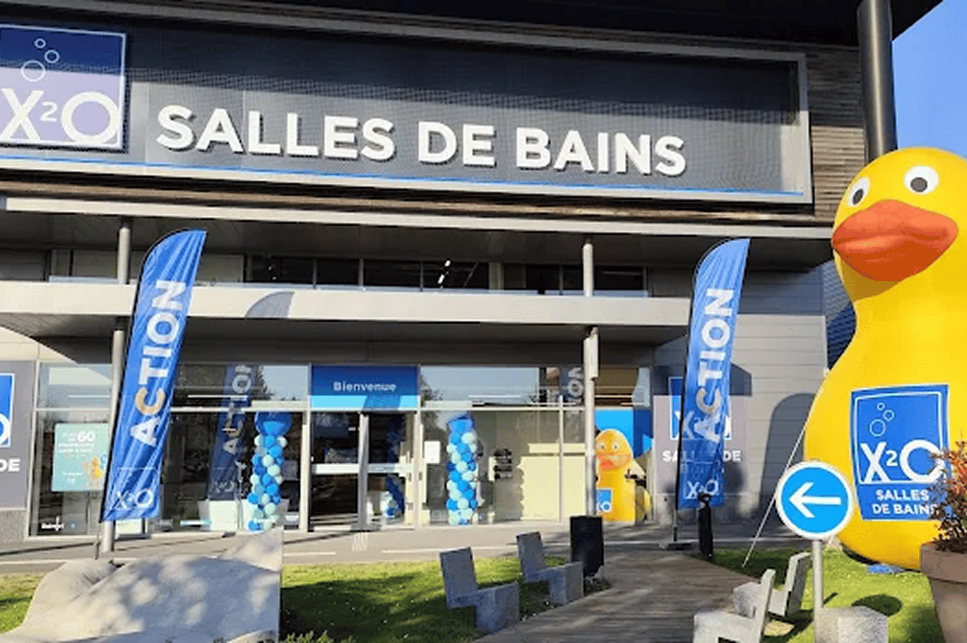 Vue extérieure d’un magasin x²o salles de bains avec façade sombre et grande inscription “SALLES DE BAINS”. Un parking avec plusieurs voitures se trouve devant, et l’entrée affiche “Bienvenue” avec des panneaux promotionnels.