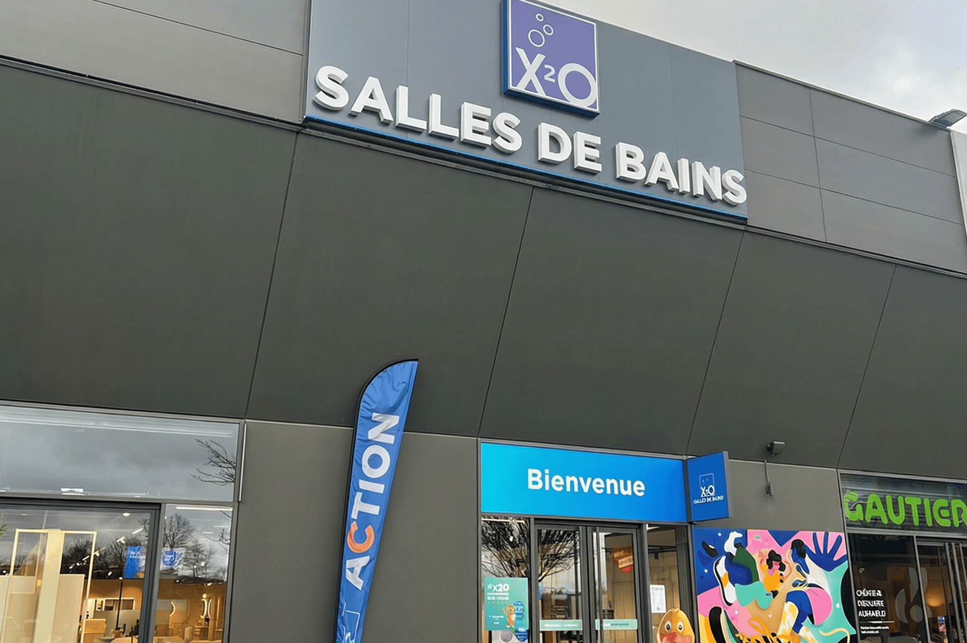 Vue extérieure d’un magasin x²o salles de bains avec une grande enseigne “X²O SALLES DE BAINS”. Une bannière “ACTION” est placée à l’entrée et le mot “Bienvenue” est visible au-dessus de la porte. Une fresque colorée et des vitrines complètent la façade dans une zone commerciale.