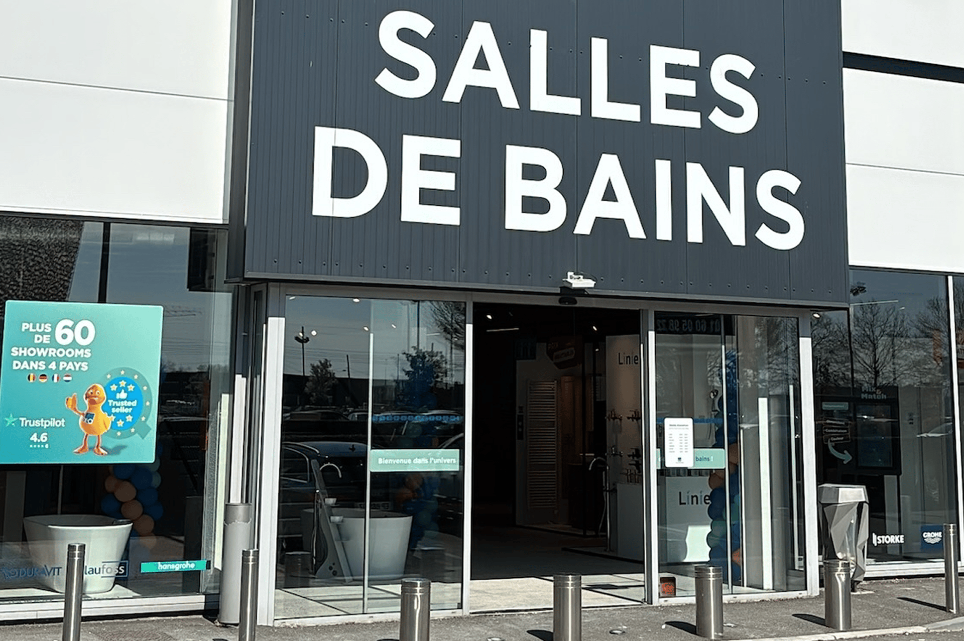 Gros plan de l’entrée d’un magasin x²o salles de bains avec l’inscription “SALLES DE BAINS” au-dessus des portes automatiques. À gauche, un panneau indique “PLUS DE 60 SHOWROOMS DANS 4 PAYS” et à droite, un grand canard jaune avec logo X²O est visible à travers la vitrine.