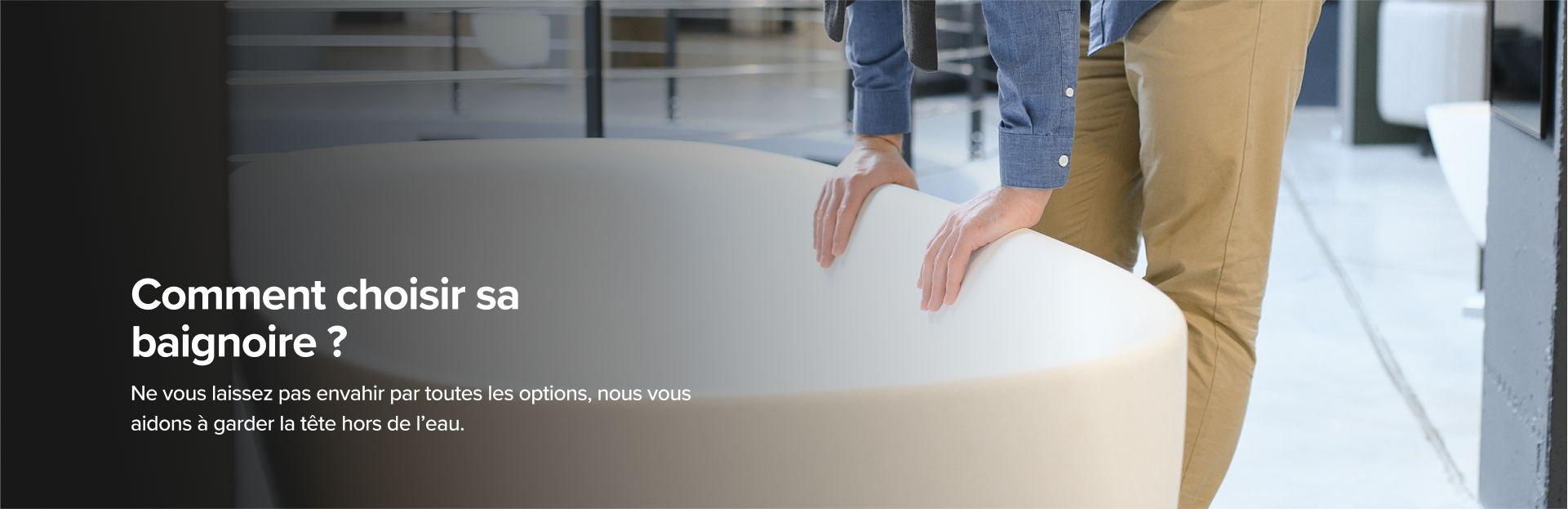 Sur cette image de x²o badkamers, un homme en pantalon beige et chemise bleue examine une grande baignoire design van x²o badkamers in een showroom met een industriële vloer en andere badmodellen op de achtergrond.