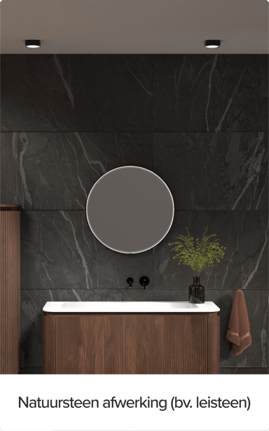 x²o badkamers meubel tegen een wand met natuursteenafwerking in leisteen-look. Rond verlicht spiegel, verticale houten kolomkasten en een zwart kraantje boven een wit werkblad benadrukken het luxe karakter.