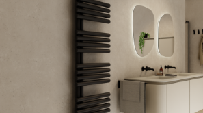 Salle de bains moderne avec un radiateur design vertical noir à lames horizontales, fixé contre un mur beige clair. À côté se trouve un meuble de salle de bains blanc avec lavabo intégré, serviette et accessoires. Deux miroirs aux angles arrondis avec éclairage indirect sont suspendus au-dessus du meuble.