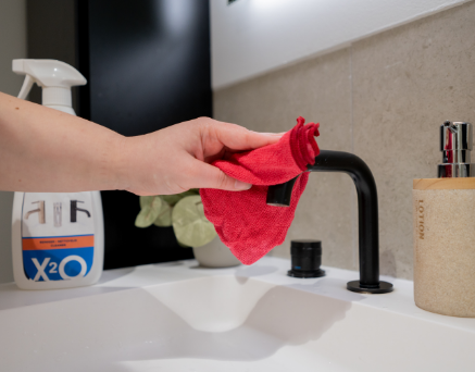 Du siehst eine Hand, die mit einem roten Tuch einen schwarzen Waschtischhahn über einem weißen Waschbecken reinigt, mit einem X²O-Reinigungsmittel im Hintergrund, als Pflegetipp von x²o Badezimmer.