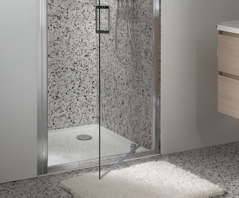 Bodengleiche Dusche mit weißer Duschwanne und transparenter Glastür vor gesprenkelten Wandfliesen. Eine weiche weiße Badematte liegt davor. Diese Dusche von x²o Badezimmer vermittelt eine saubere und frische Badatmosphäre.