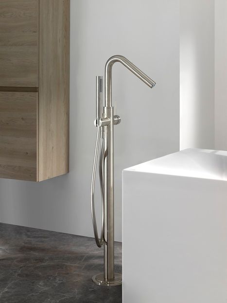 Detailfoto van een vrijstaande inoxlook badkraan in coolmetal afwerking naast een strak wit ligbad. De kraan staat op een donkere vloer en wordt gecombineerd met een houten badkamerkast op de achtergrond. Deze moderne en minimalistische badkameropstelling toont de stijlvolle kranen van x²o badkamers.