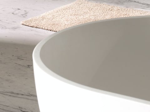 Detail van een vrijstaand bad met afgeronde rand, geplaatst op een marmeren vloer en een beige badmat.