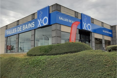 x²o badkamers showroom in Charleroi met blauwe luifel, glazen gevel en toegangsdeuren met zicht op interieur.