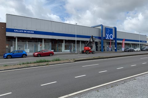 Grand showroom x²o salles de bains à Liège le long d’une route avec façade vitrée, signalisation bleue et voitures stationnées sous un ciel nuageux.