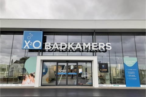 Façade d’entrée du showroom x²o salles de bains à Ostende avec logo “BADKAMERS”, portes vitrées automatiques et affiches promotionnelles.