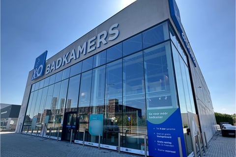 Showroom moderne x²o salles de bains à Audenarde avec façade vitrée, branding bleue et parking à l’avant.