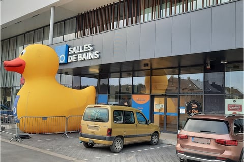 Showroom x²o salles de bains à Soignies avec un grand canard jaune décoratif devant le bâtiment et voitures stationnées.