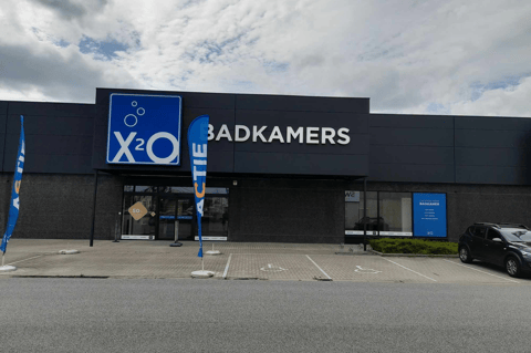 Magasin X²O salles de bains avec façade sombre, entrée centrale et drapeau promotionnel devant le bâtiment – X²O salles de bains