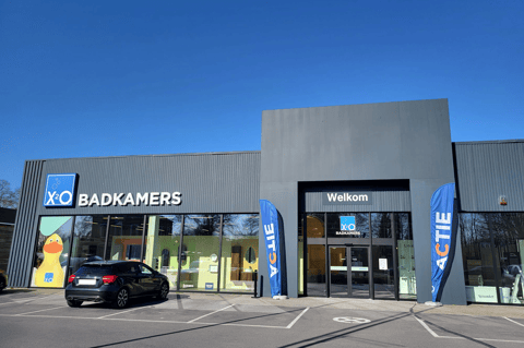 Showroom moderne X²O salles de bains avec entrée accueillante, façade vitrée et signalisation “Welkom”, complété par des drapeaux promotionnels et un parking spacieux – X²O salles de bains