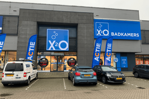 Gevel van X²O Badkamers winkel met parking en promotiebanners voor de ingang – X²O badkamers