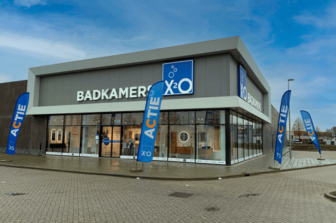 Moderne X²O Badkamers showroom op hoeklocatie met grote glaspartijen, duidelijke branding en opvallende actie-vlaggen die klanten uitnodigen om binnen inspiratie op te doen – X²O badkamers
