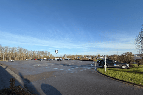 Grand parking presque vide avec quelques voitures sur les côtés, sous un ciel bleu clair et des arbres en arrière-plan.