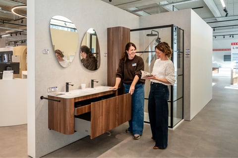 In diesem Showroom von x²o Badezimmer berät dich eine Mitarbeiterin gemeinsam mit einer Kundin vor einem Holz-Badezimmermöbel mit Doppelwaschbecken, wobei eine Schublade geöffnet ist. Darüber hängen zwei organisch geformte Spiegel und im Hintergrund siehst du eine Glasdusche mit schwarzen Profilen in einem hellen, modernen Raum.