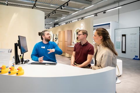 In einem Showroom von x²o Badezimmer erklärt dir ein Mitarbeiter einem Paar an einem runden Beratungstresen mit Computer verschiedene Optionen. Auf dem Tresen stehen gelbe Badeenten als Dekoration. Im Hintergrund siehst du mehrere Badezimmerausstellungen mit Spiegeln, Möbeln und Beleuchtung.
