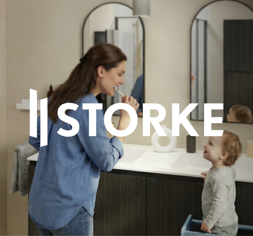 Vrouw die haar tanden poetst naast een jong kind aan een dubbele wastafel met geribbeld donker badkamermeubel en twee rechthoekige spiegels, in een lichte badkamer met raambekleding en accessoires, afgebeeld als sfeerbeeld voor Storke bij x²o badkamers.