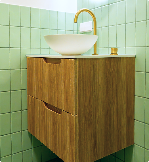 In deze x²o badkamers zie je een houten wastafelmeubel met een witte waskom en een hoge, gouden kraan, geplaatst tegen een wand met mintgroene tegels.