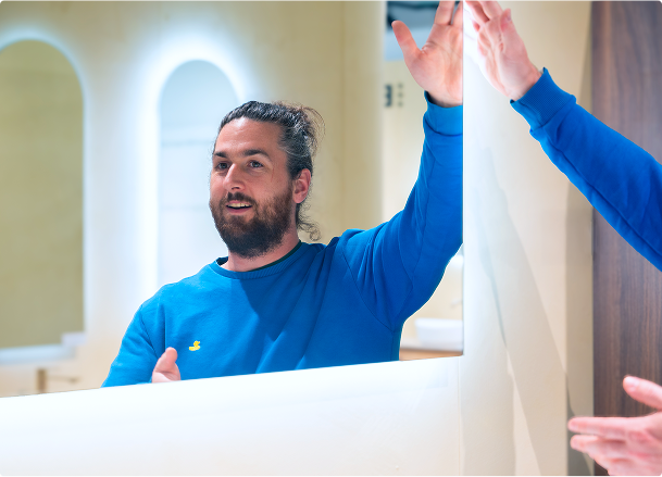Een man in een blauwe trui met een geborduurd eendje test een grote badkamerspiegel met geïntegreerde verlichting in de showroom van X²O badkamers.