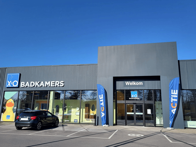 Strakke X²O Badkamers showroom met moderne gevel, duidelijke “Welkom”-ingang en opvallende actie-vlaggen, met zicht op inspirerende badkameropstellingen binnen – X²O badkamers