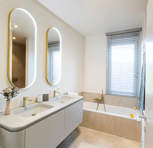Salle de bains x²o lumineuse et fraîche avec une disposition moderne. À gauche, meuble suspendu avec double vasque, robinets dorés et deux miroirs verticaux à éclairage LED. À droite, une baignoire encastrée avec robinet assorti et une fenêtre avec des stores laisse entrer la lumière naturelle.