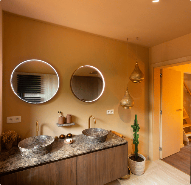 Ambiance orientale chaleureuse dans cette salle de bains x²o avec des tons terre, deux miroirs ronds à éclairage intégré et un meuble lavabo double en bois avec plan de travail effet marbre. Des lanternes suspendues et un cactus en pot tressé sont visibles près de la porte.