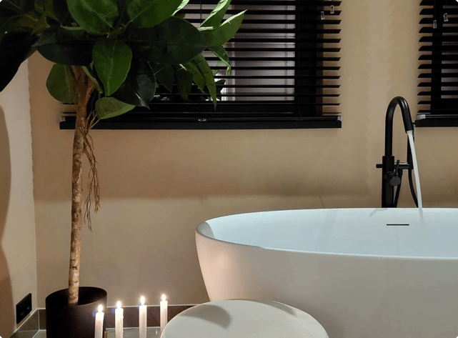 Salle de bains x²o chaleureuse avec baignoire blanche îlot, robinet noir sur pied et ambiance tamisée. Six bougies allumées et une grande plante verte ajoutent une touche relaxante. En arrière-plan, des stores noirs couvrent la fenêtre.
