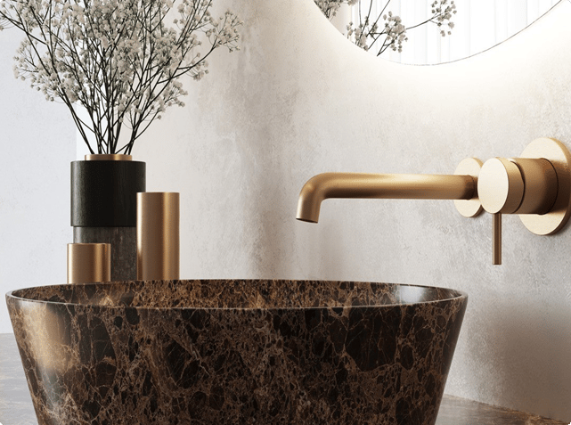 Détail d’un lavabo luxueux dans une salle de bains x²o avec vasque effet marbre foncé et robinet mural doré. À gauche, des éléments décoratifs cylindriques dorés et noirs accompagnés de fleurs blanches délicates. Le tout dans des tons doux et naturels.