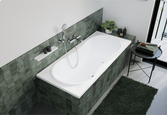Deze foto van x²o badkamers toont een ingebouwde witte badkuip in een betegelde ombouw met groene tegels, een chromen douchekop en kraan, een plantje en zwarte accessoires op de rand en een ronde zwarte bijzettafel met handdoek en borstel.