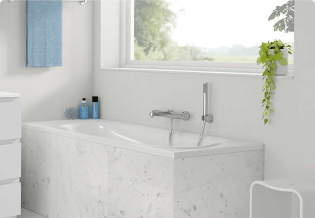 Op deze foto van x²o badkamers zie je een kleine witte ingebouwde badkuip met een witte marmeren ombouw, een chromen wandkraan met handdouche, twee blauwe flessen en een sponsje op de badrand, een blauwe handdoek aan de muur en een hangplant op de vensterbank met vrij uitzicht naar buiten.