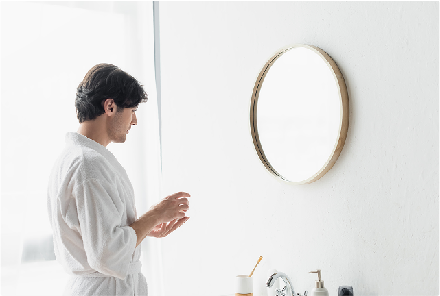 Homme en peignoir blanc devant un miroir de salle de bains rond à bord doré, dans une salle de bains lumineuse – routine quotidienne chez x²o salles de bains.